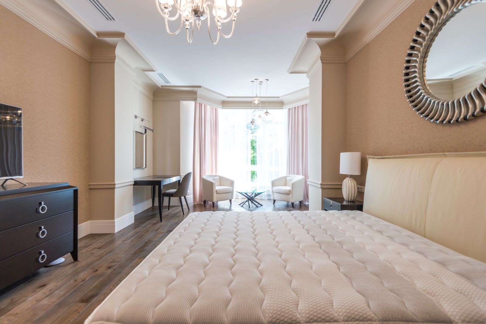 Spacious classic bedroom with a quilted cream bed, a warm taupe wall finish and a sitting area by the window