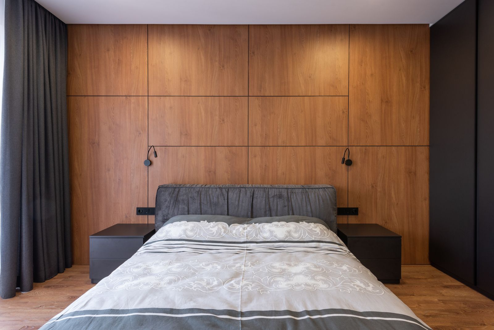 Master bedroom with a full-height timber wall, a grey upholstered bed with reading lights and dark curtains