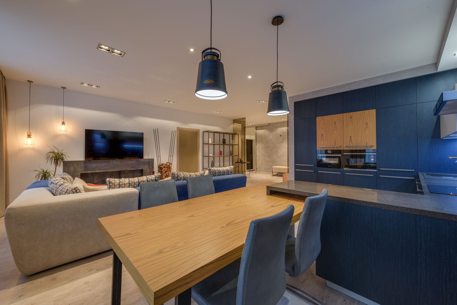 Open-plan home with a deep blue kitchen, a timber dining table with blue chairs and a lounge with pendant lights