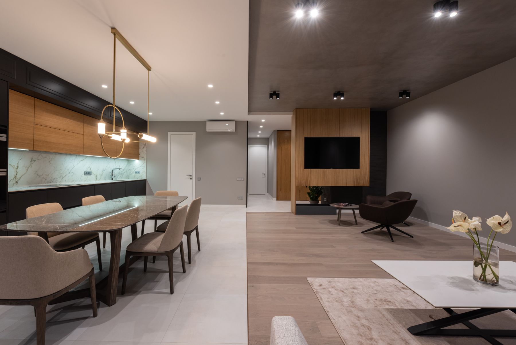 Open-plan home interior with a long dining table beneath slim pendant lights, a kitchen with timber cabinetry and a lounge beyond