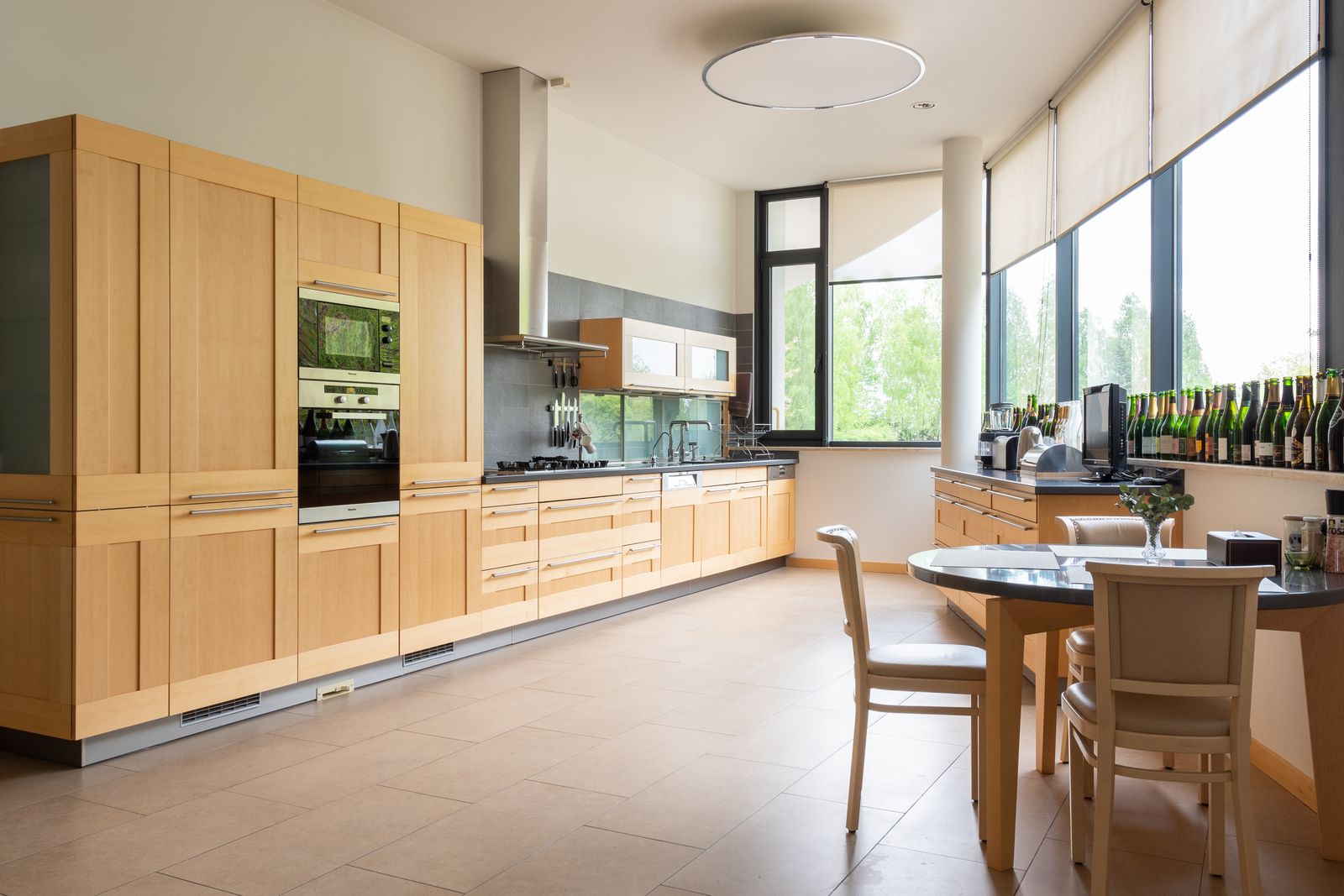 Long modular kitchen with oak cabinetry running along a wall of windows and a round dining table