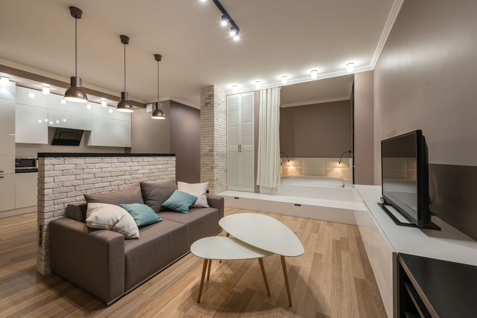 Compact open-plan living area with a taupe sofa, a brick-effect partition and pendant lighting over a kitchenette