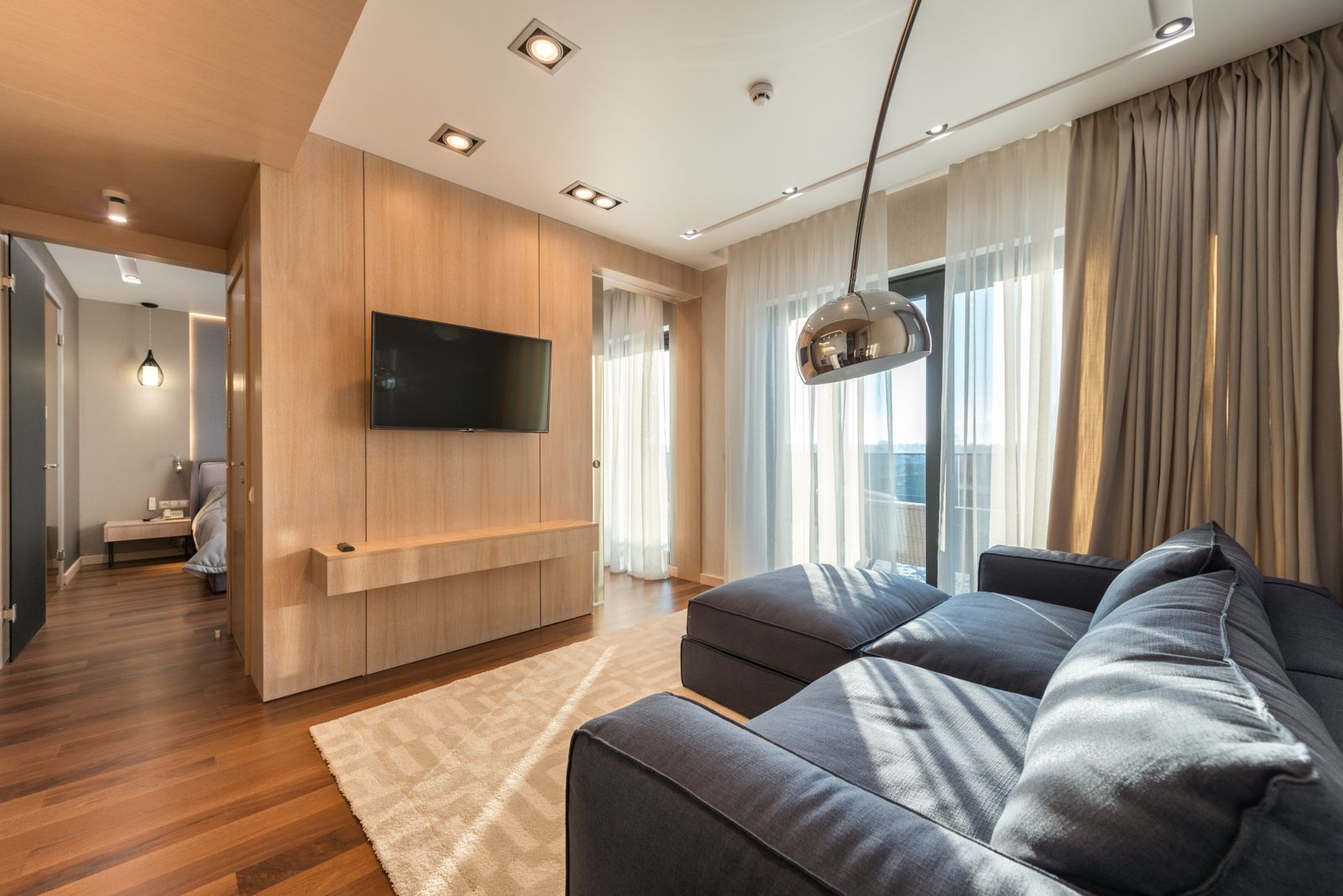 Living room with a charcoal leather sofa, a light timber media wall and an arched floor lamp beside sheer curtains
