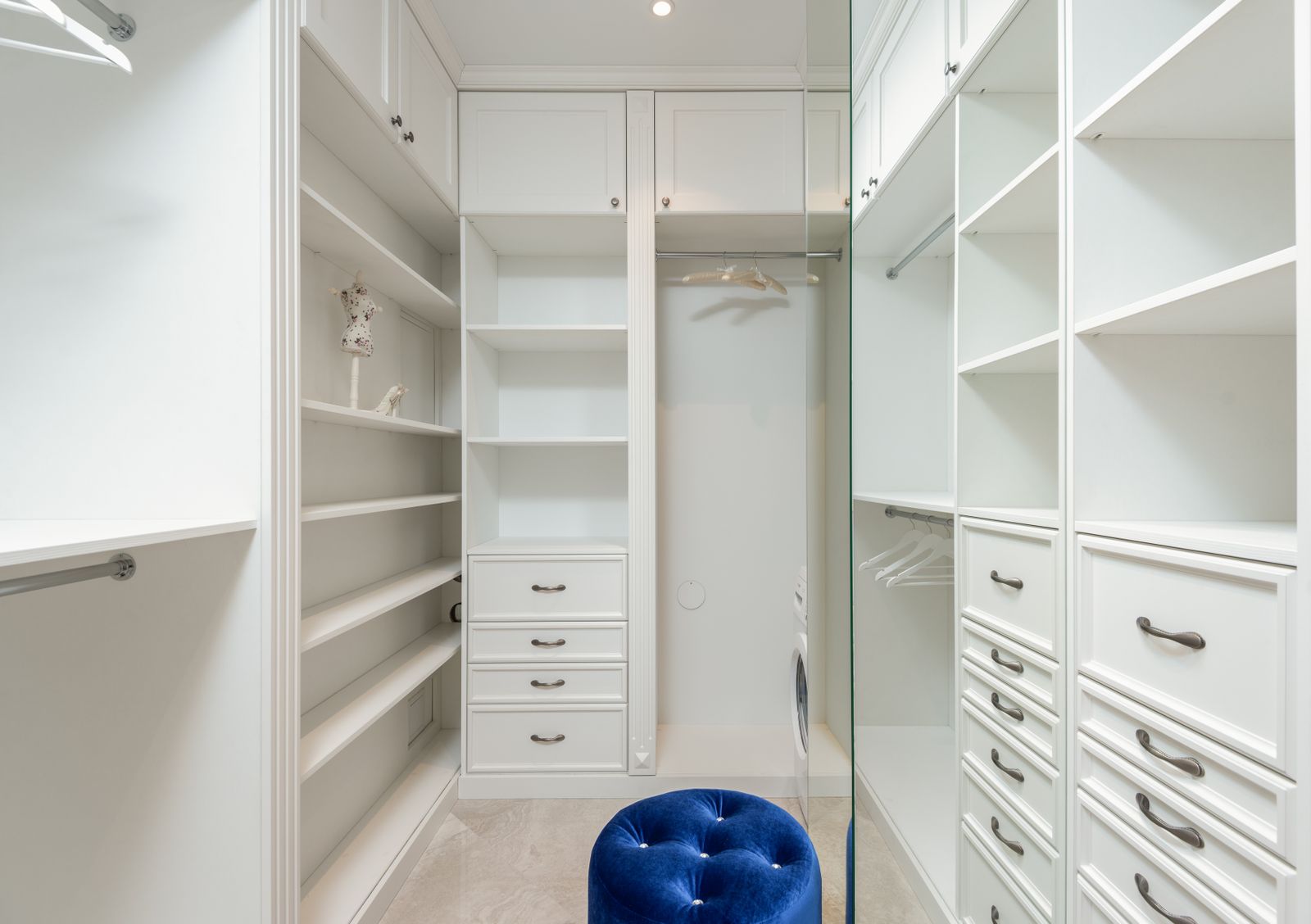 White panelled walk-in wardrobe with drawers, shelving, hanging space and a blue upholstered pouffe